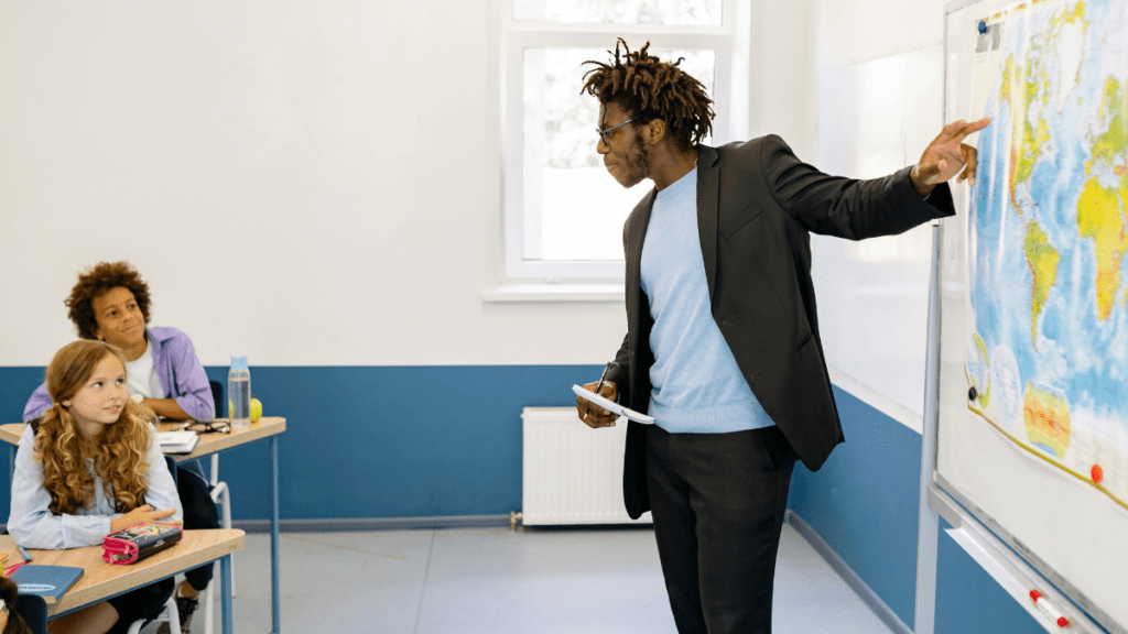 A male teacher in front of a classroom pointing to a map while students are looking towards the map - using options to connect one of the strategies for teaching current events