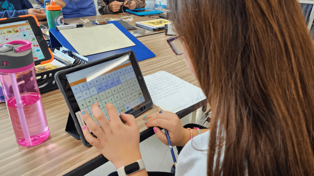 high school girl seated at a classroom table using an AAC device.